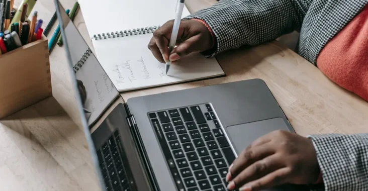 Pessoa em mesa de escritório com notebook aberto e caderno com anotações, enquanto escreve com caneta e usa o computador, ilustrando como o curso de Administração ajuda a preparar futuros líderes